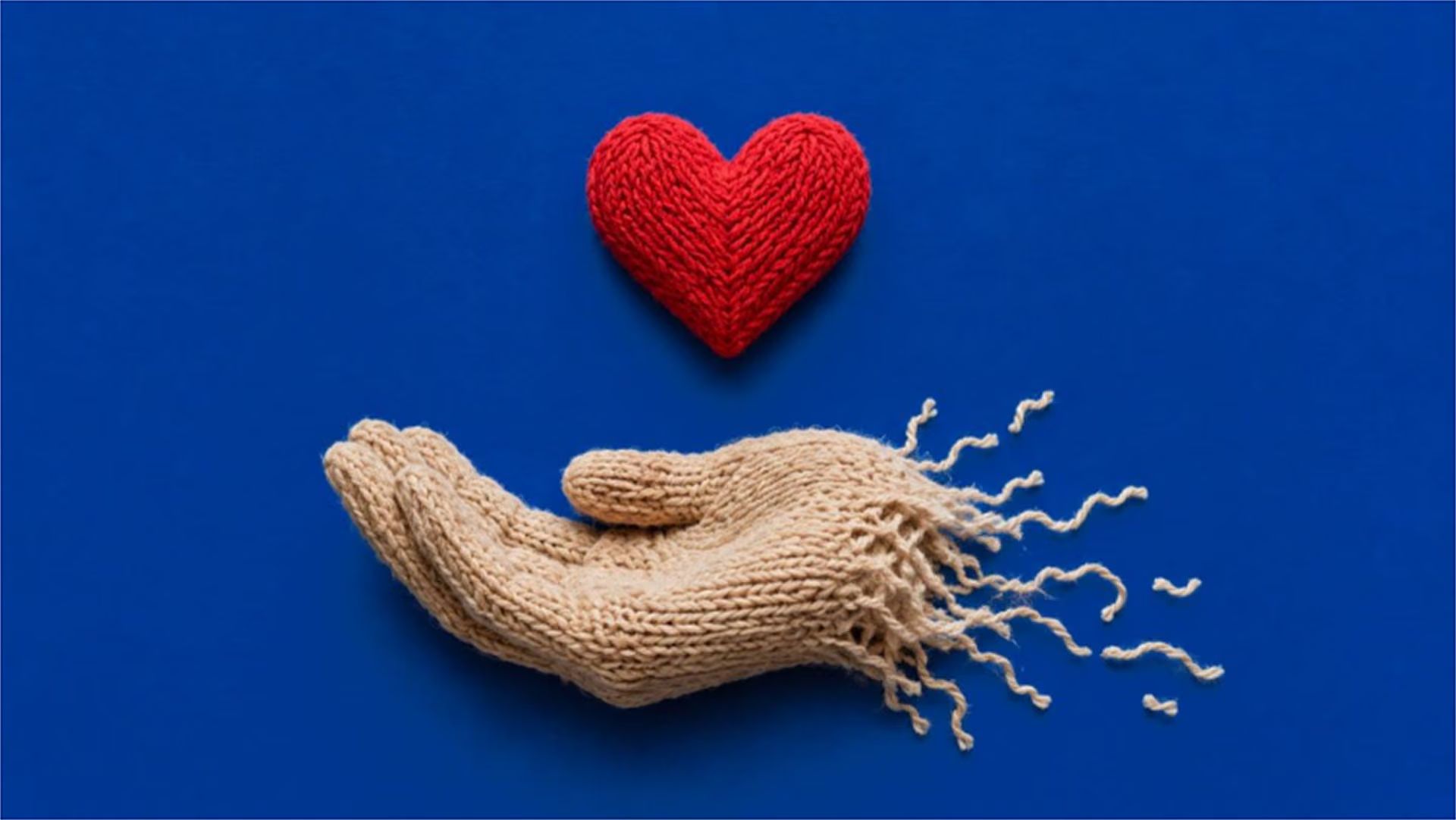A knitted red heart resting above a knitted hand, unravelling at the wrist.