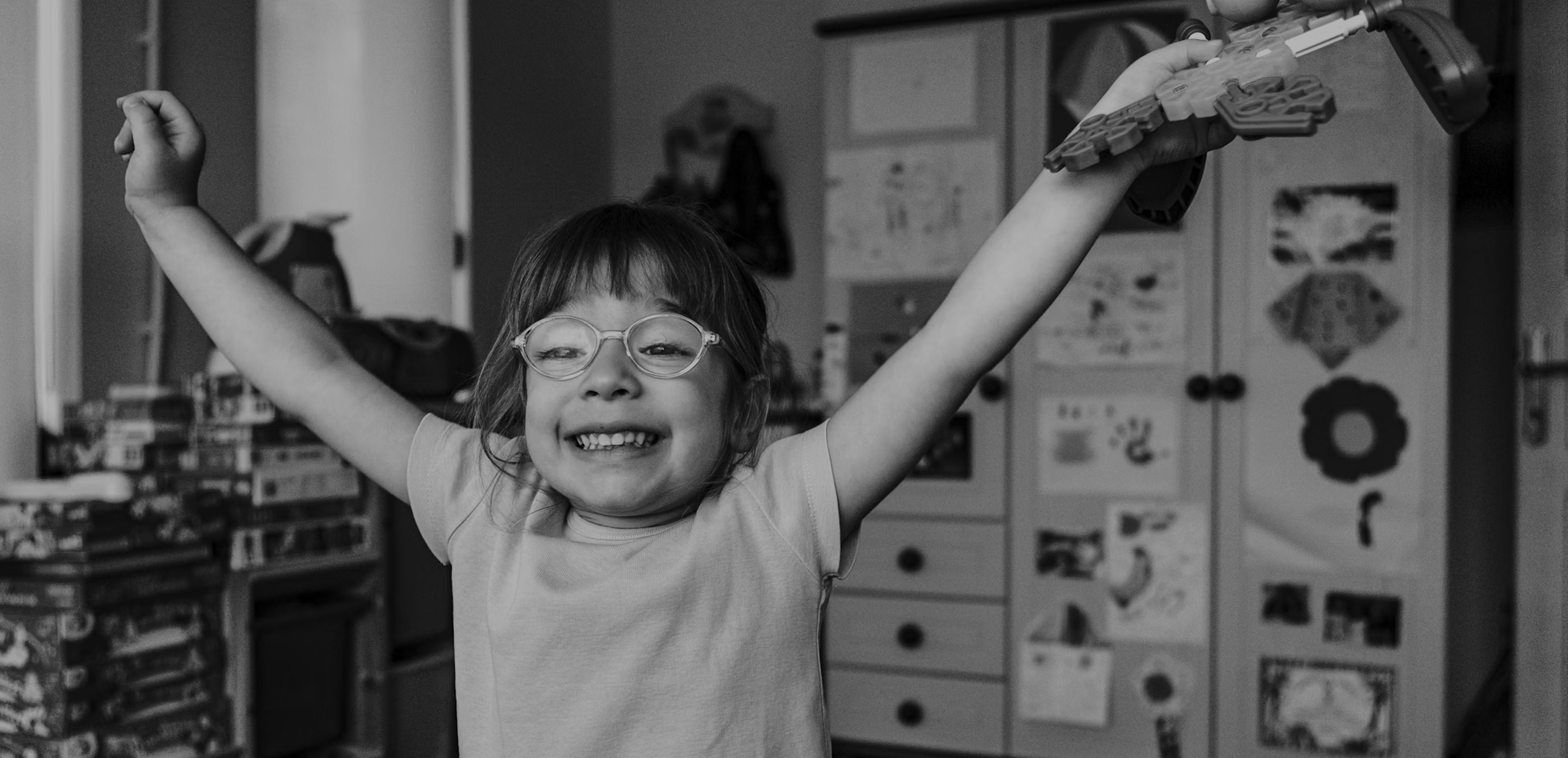 A girl in glasses grinning with both arms flung up, in a playroom.